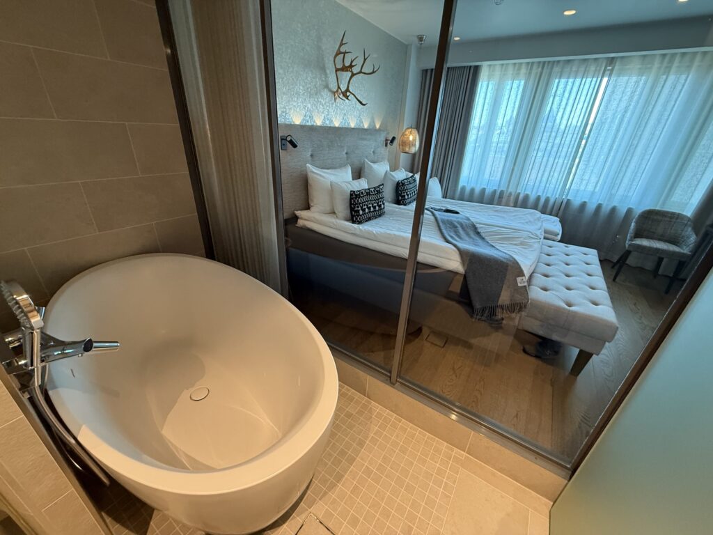 A modern hotel room with a large white bathtub in the bathroom, looking through a glass wall into the bedroom where a bed is decorated with deer antlers above the headboard.