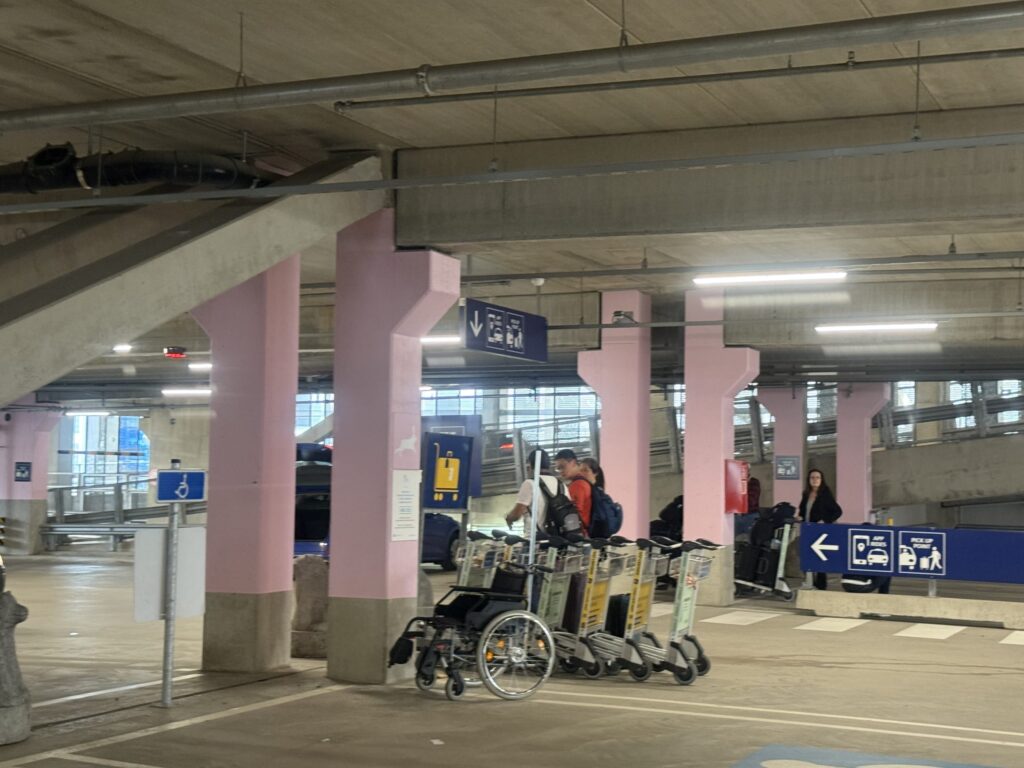 An Uber pickup point in an airport parking garage, featuring striking pink pillars. A few travelers, luggage carts, and a wheelchair are visible.