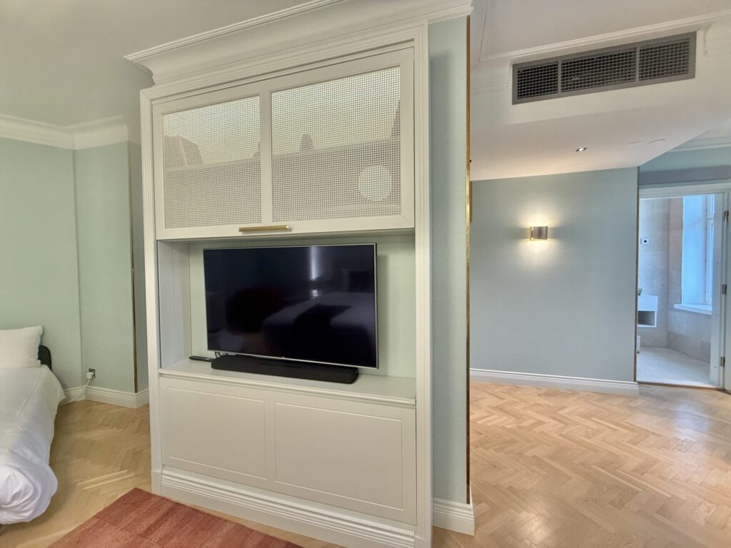 A white, built-in TV cabinet installed in a room with pale blue walls. A television is housed within the cabinet, and to the right, a hallway with herringbone flooring and the entrance to a bathroom beyond it are visible.