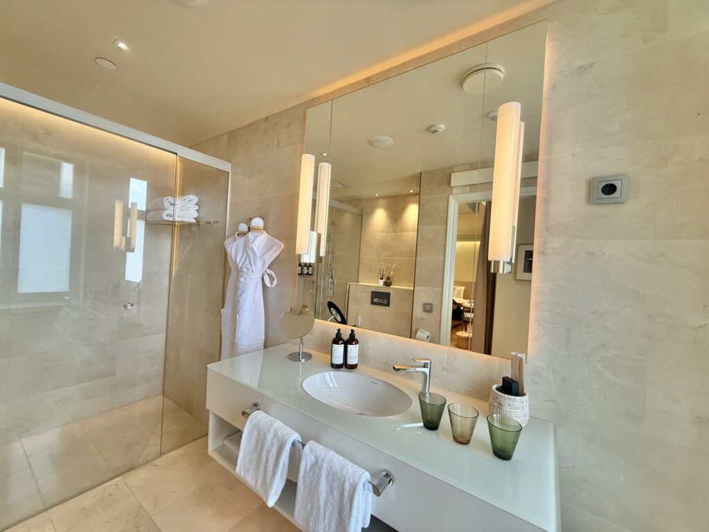A modern, luxurious bathroom with a beige color scheme. A white vanity holds stylish glasses, with a large mirror on the wall and a glass-walled shower room to the side.