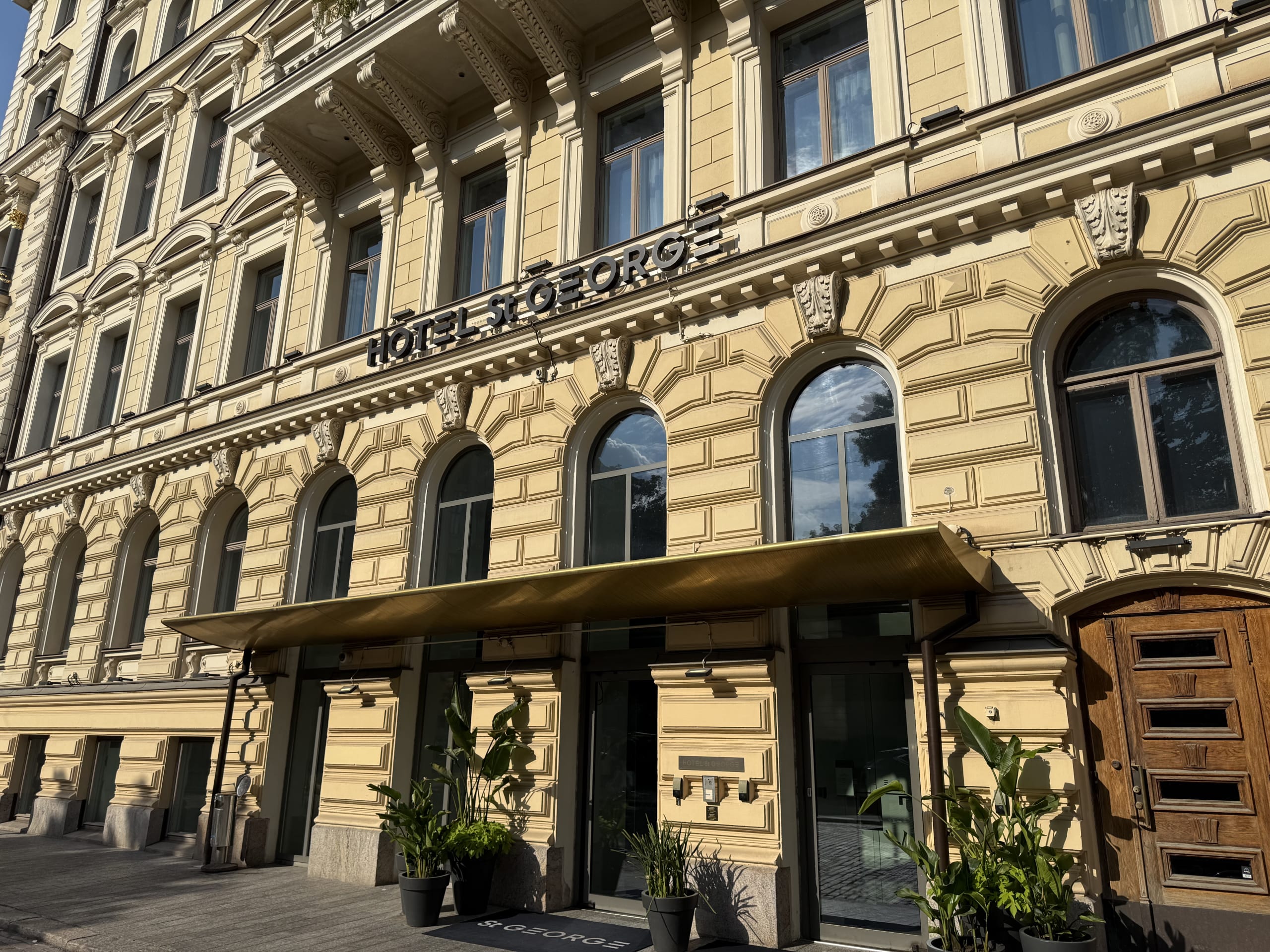 The sunlit, cream-yellow facade of the historic Hotel St. George in Helsinki, showing the main entrance under a modern golden canopy.
