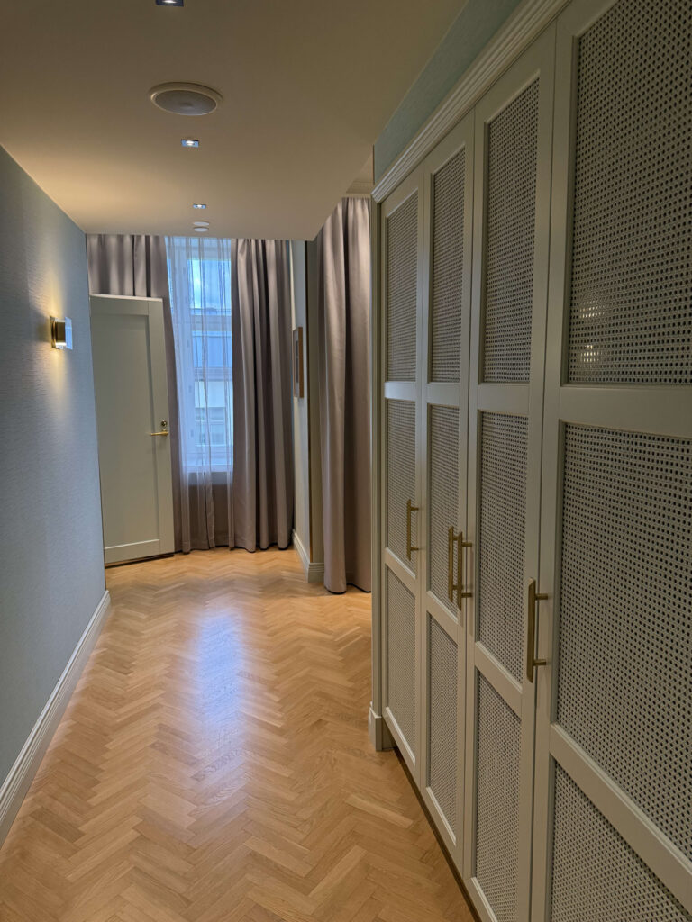 An elegant hallway with a herringbone pattern wood floor. On the right are closets with cane webbing doors, and on the left, a modern light fixture illuminates a light blue wall.