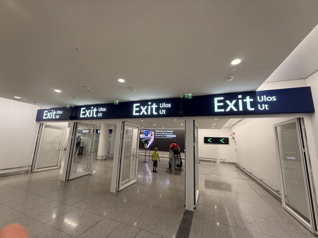 ヘルシンキ空港の到着ロビーにある出口ゲート。英語、フィンランド語、スウェーデン語で「Exit」と書かれた案内標識が複数あり、数人の旅行者が出口に向かっている。