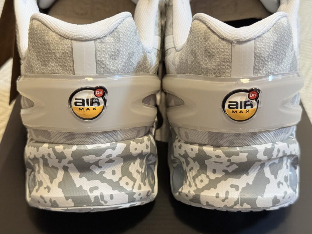 Close-up of the heels of a pair of white and grey sneakers, showing the grey camouflage-patterned midsole and the 3D Air Max Dn logos on the heel counters.