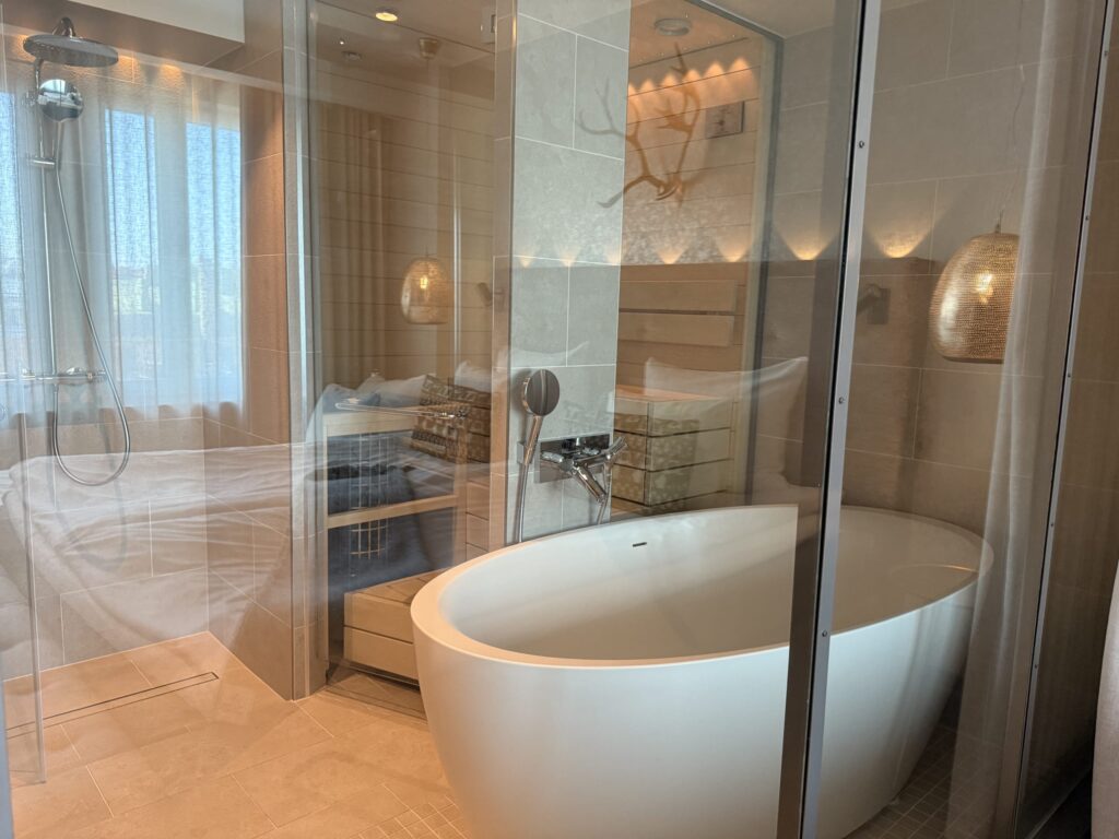 A modern bathroom with beige tiles, featuring a white freestanding bathtub in the foreground and a glass-enclosed shower and private wooden sauna in the background.