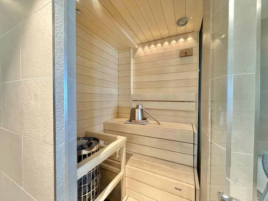 The interior of a modern home sauna made of light-colored wood. It features a two-tiered bench, a sauna stove, and a bucket and ladle for löyly.