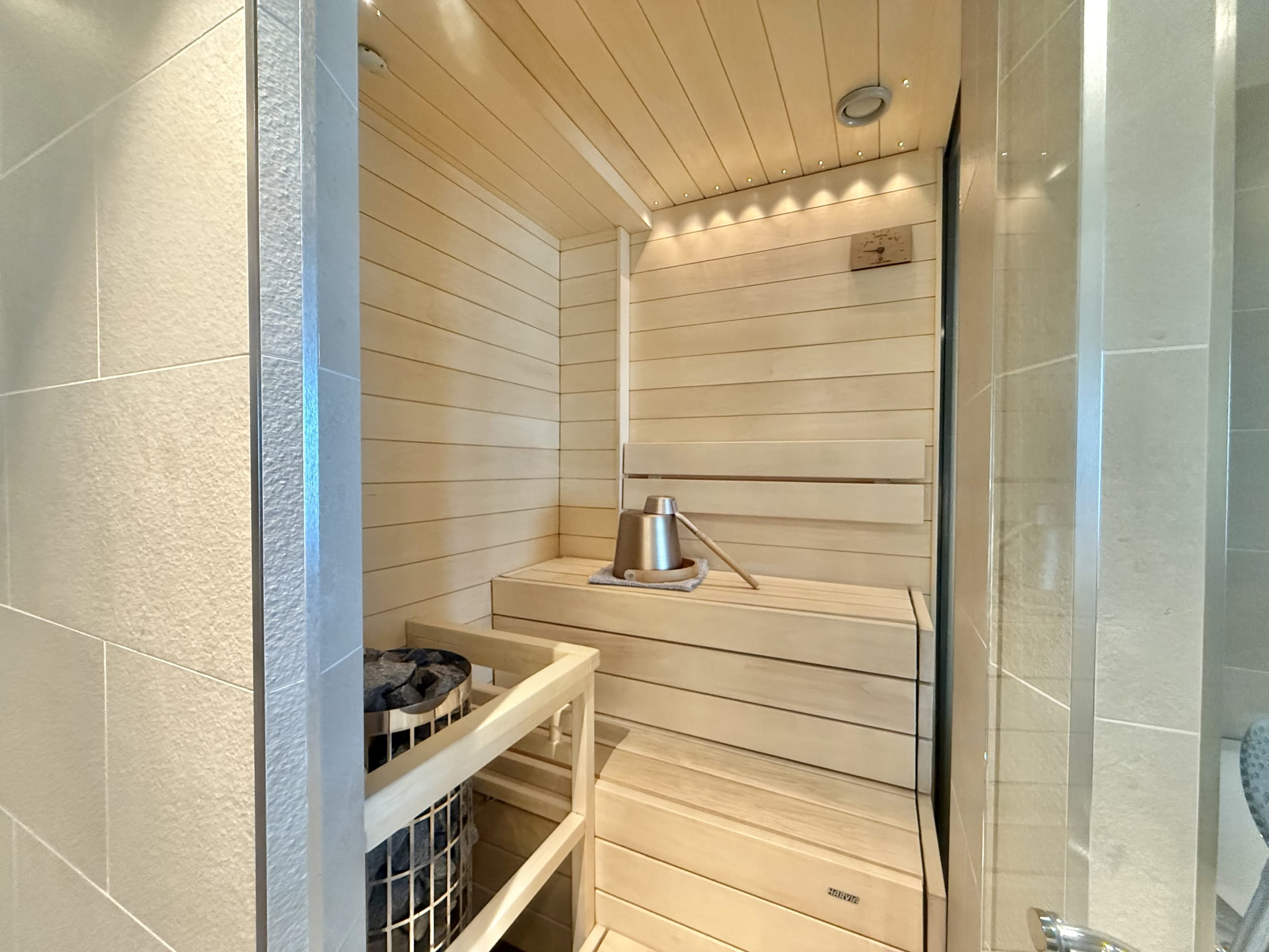 The interior of a modern home sauna made of light-colored wood. It features a two-tiered bench, a sauna stove, and a bucket and ladle for löyly.