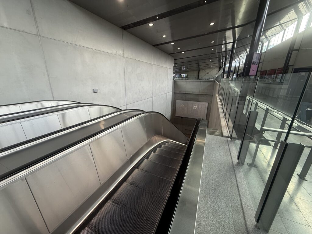 View from the top of an escalator heading down in a modern station interior. There is a concrete wall on the left, glass panels on the right, and the area is lit by ceiling lights and light from windows.