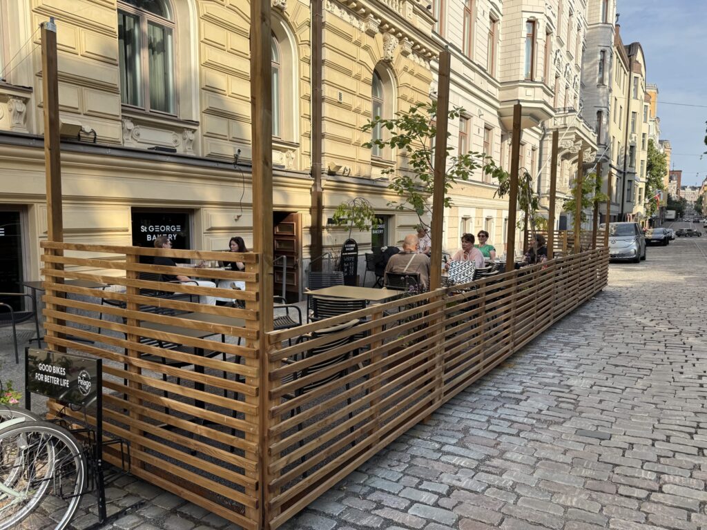 The outdoor terrace seating of St. George Bakery on a cobblestone street in Helsinki. People are relaxing and chatting at tables enclosed by a wooden fence.