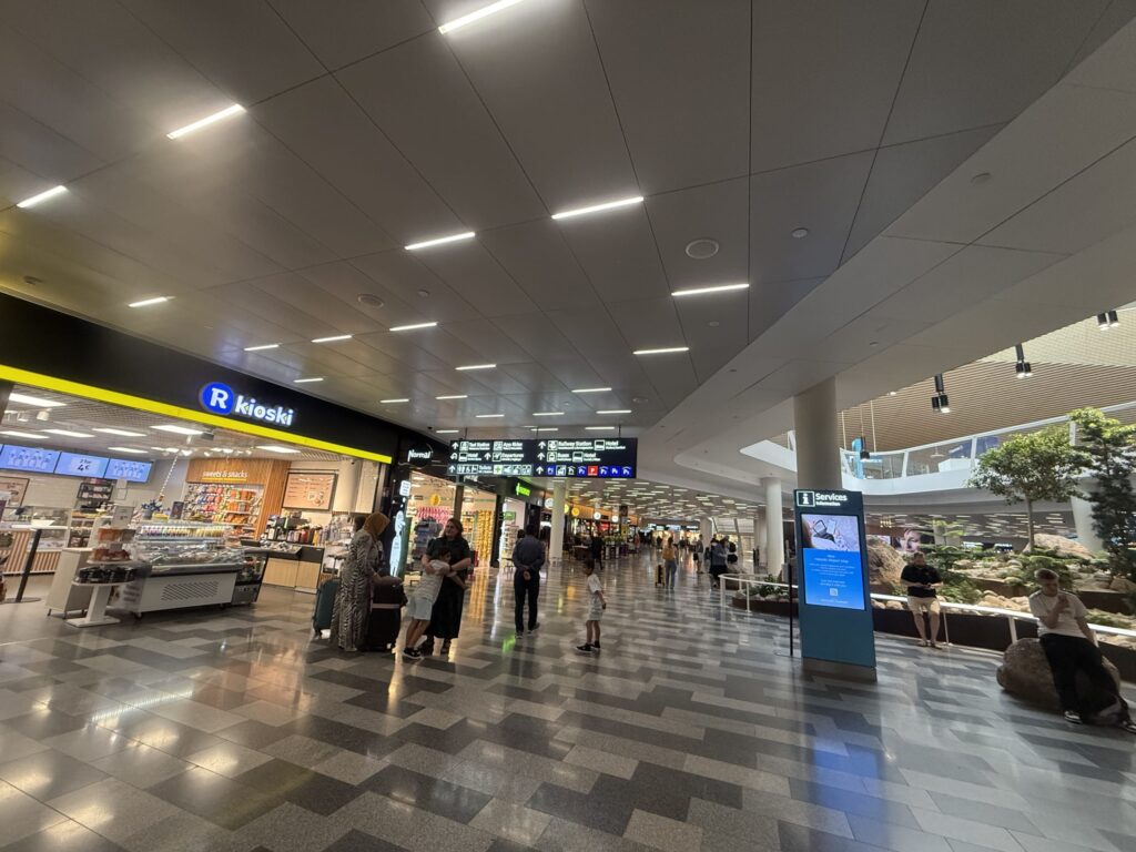 The modern arrivals hall of Helsinki-Vantaa Airport, with an R-kioski store on the left and travelers walking by.
