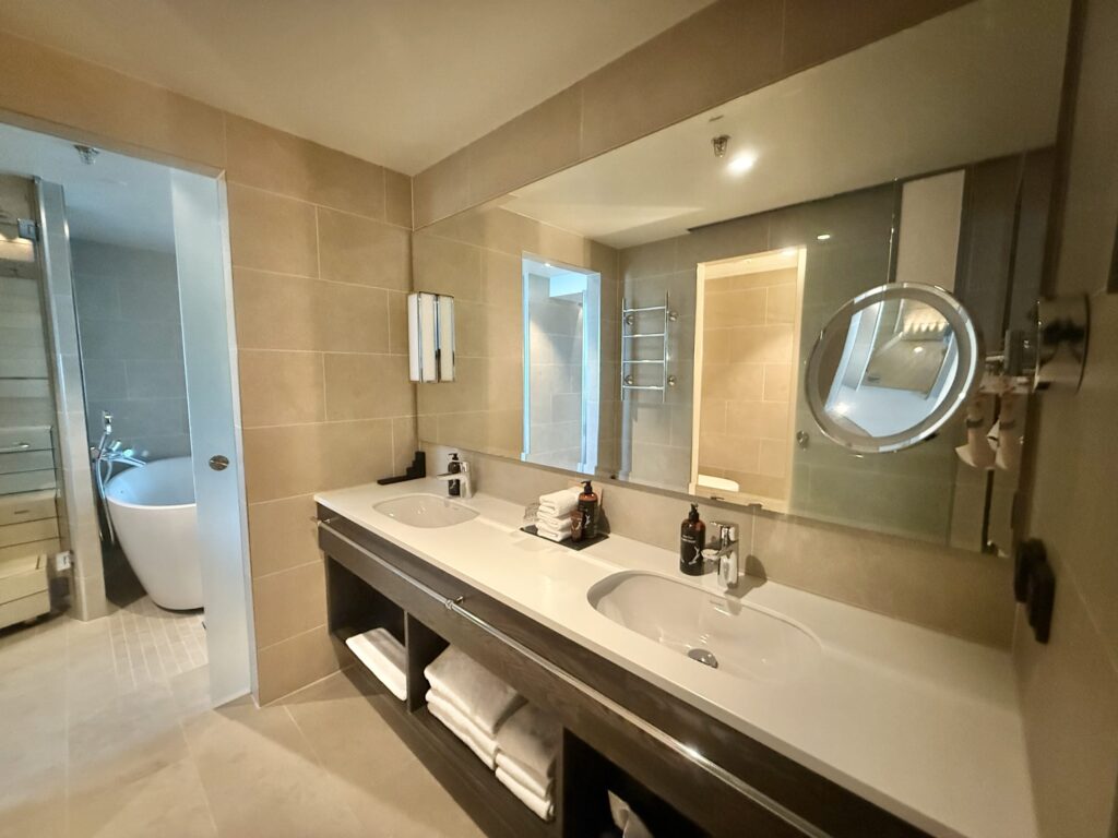 A modern bathroom vanity in a hotel suite, featuring two sinks, a large mirror, and a view of the bathtub in the adjacent room.