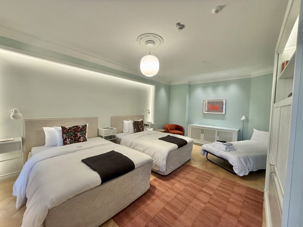 A spacious hotel room with pastel green walls, featuring two twin beds, an extra rollaway bed, and modern Scandinavian-style furniture.