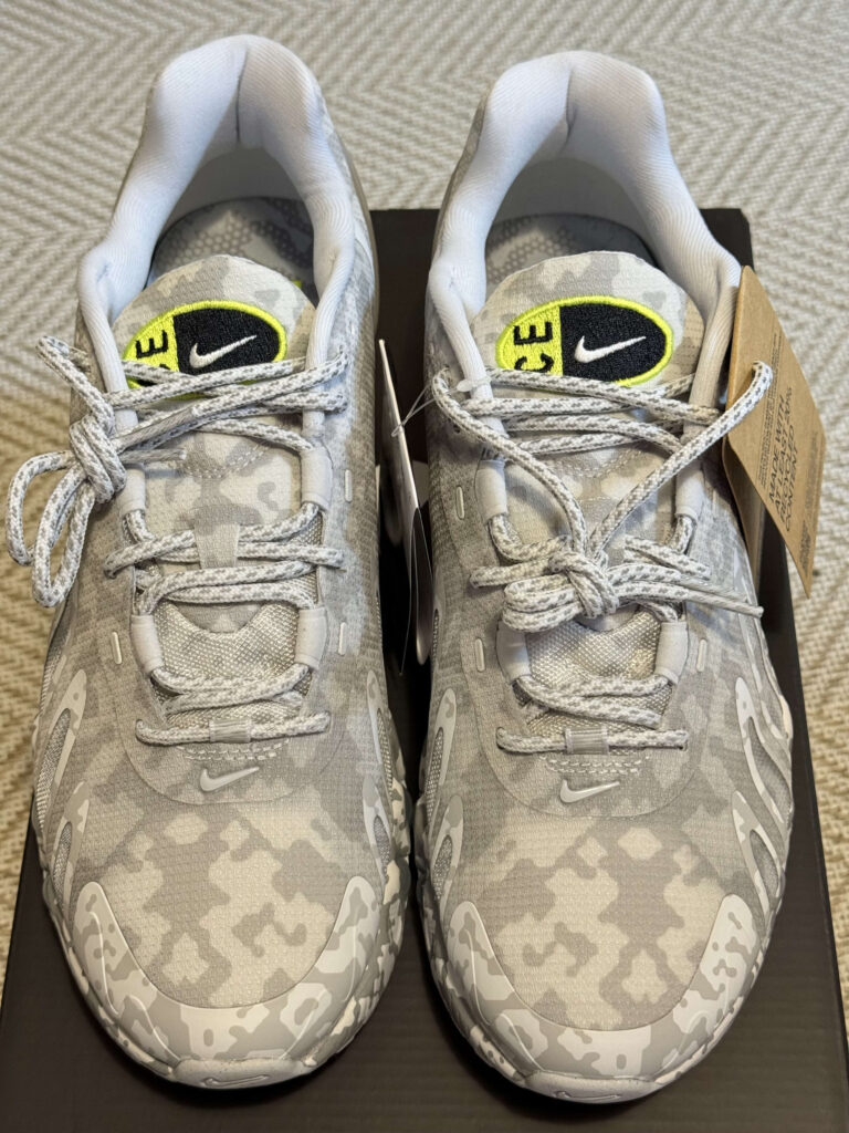 A pair of Nike sneakers with a grey camouflage pattern, shot from the front, showing the yellow logo on the tongue and the patterned laces.