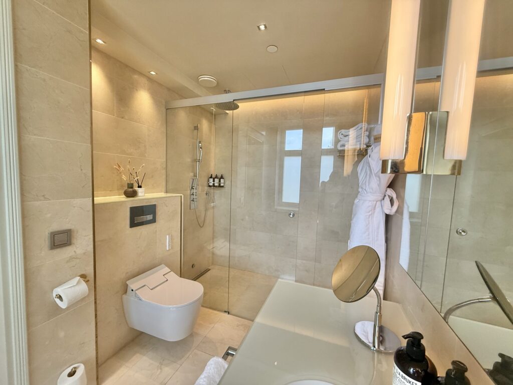 A modern bathroom with beige tiles, featuring a glass-enclosed walk-in shower and a wall-mounted toilet.