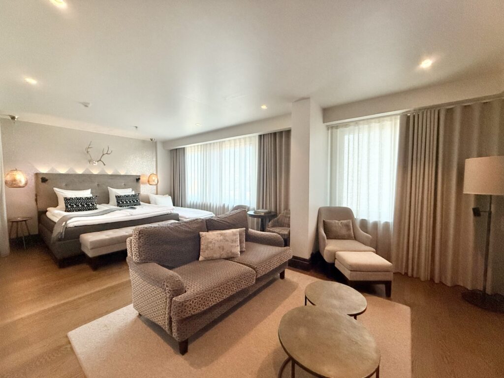 A spacious hotel room decorated in natural tones. A bed is in the background, with a sofa and lounge chair arranged in the foreground.