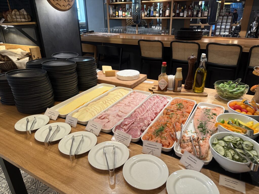 A rich breakfast buffet spread on a restaurant's wooden counter, featuring smoked salmon, cured meats, cheeses, and fresh vegetables.