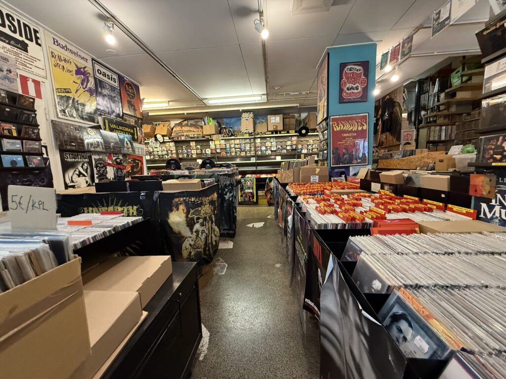 The interior of a record store filled to the brim with vinyl records. Bins of records line a narrow aisle, and the walls are decorated with posters of bands.