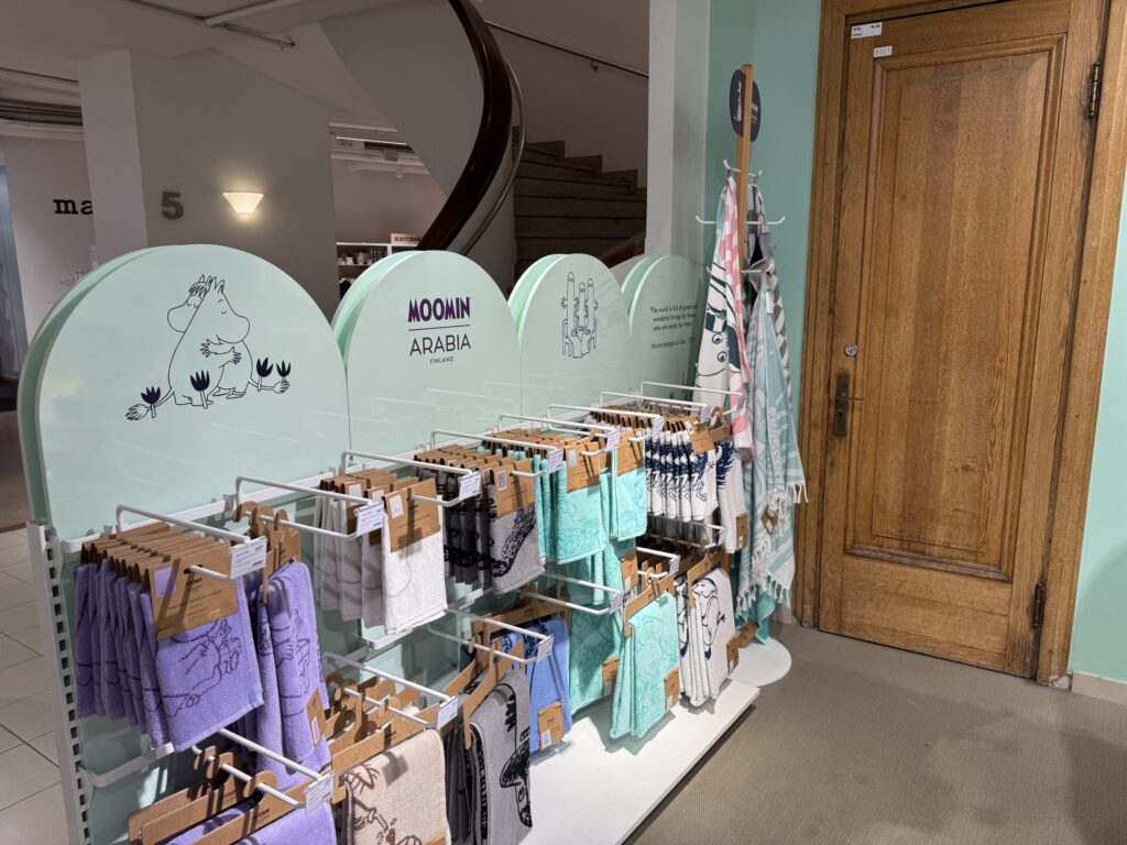 An in-store display of Moomin Arabia towels. Various towels featuring Moomin characters hang on a white rack with a mint green backdrop, next to an old-fashioned wooden door.