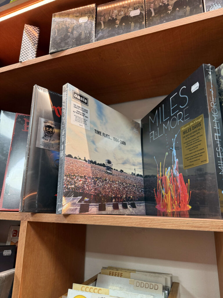 Vinyl records by Oasis, Motörhead, and Miles Davis are displayed on a wooden shelf in a record store. In the center, Oasis's 'Time Flies... 1994-2009' is facing forward.