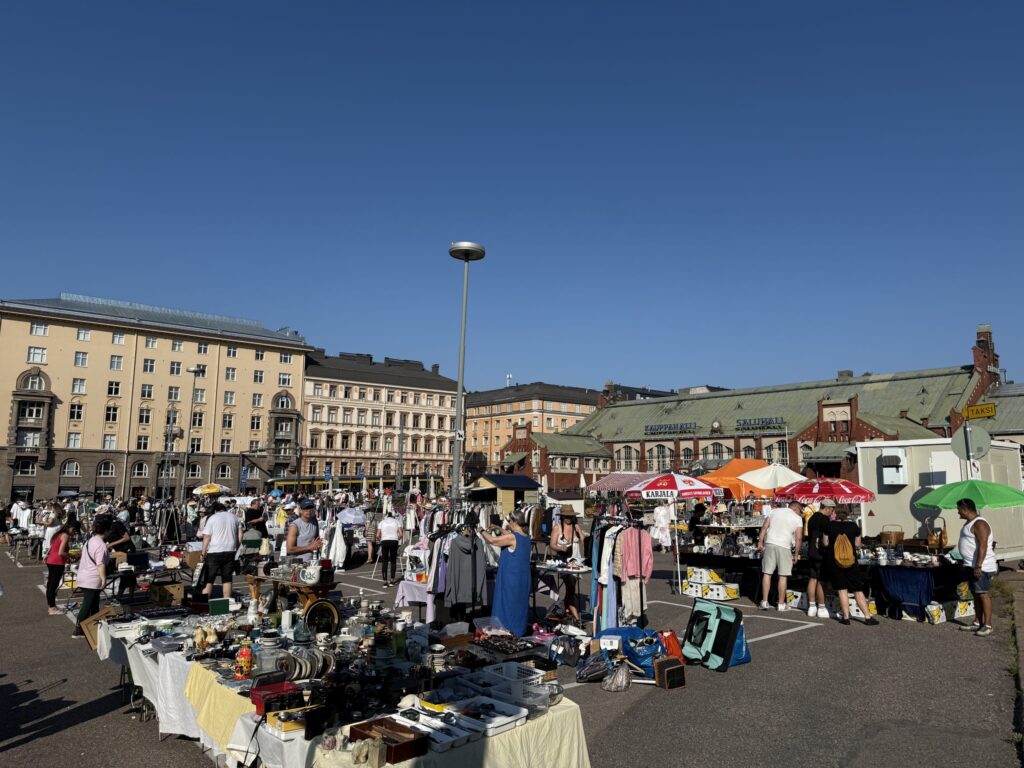 The Hietalahti flea market on a sunny day, with many people browsing stalls selling second-hand clothes, dishes, and other goods.