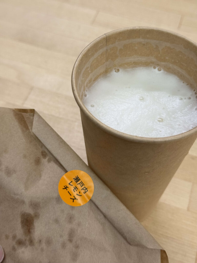 A paper cup of beer and a curry bread in a paper bag with a sticker that reads 'Setouchi Lemon Cheese' in Japanese, placed on a wood-grain table.