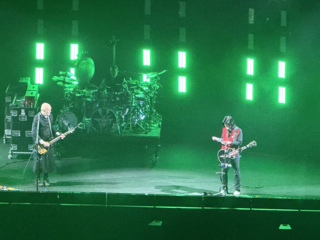 The Smashing Pumpkins performing on a stage illuminated by green lights. Billy Corgan is on the left, James Iha is on the right, and the drummer is visible in the background.