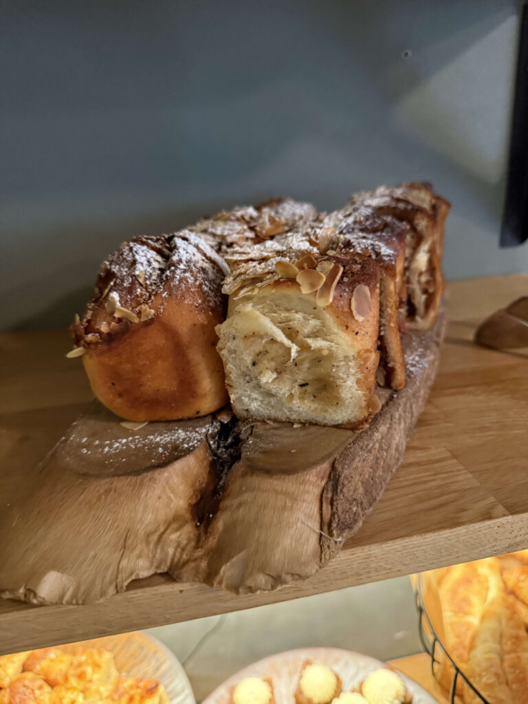 A sweet bread loaf topped with sliced almonds and powdered sugar, resting on a wooden board. A portion of the bread is cut, showing the fluffy, layered texture inside.