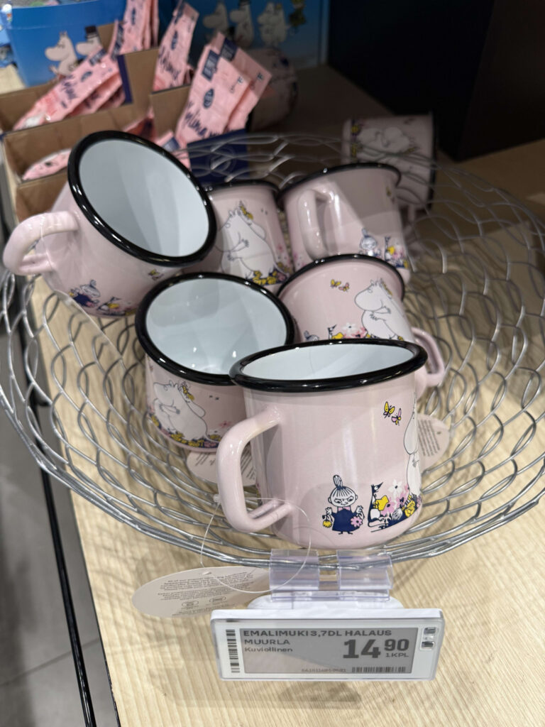 Pink enamel mugs featuring Moomin characters displayed in a wire basket at a store.