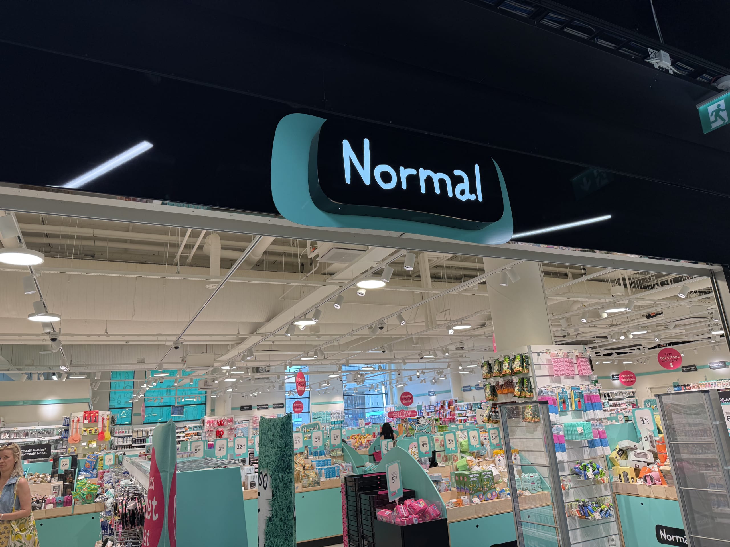 The entrance to a 'Normal' variety store, featuring its signature black sign with a turquoise border. The bright interior is visible, with shelves packed with products.