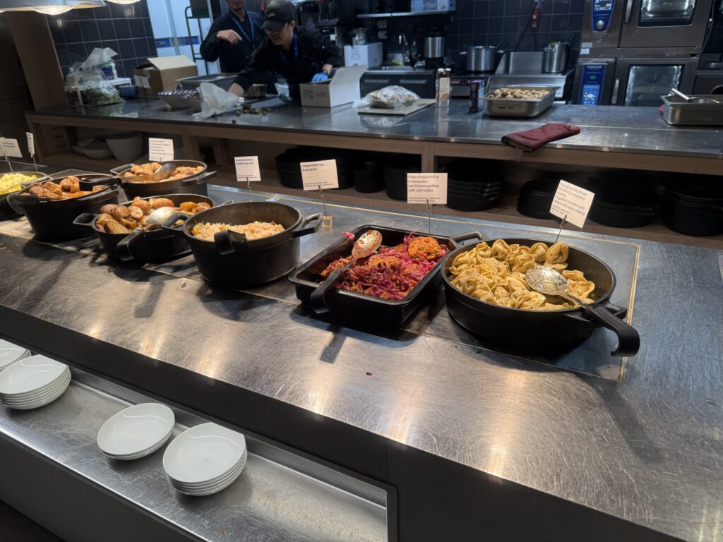 A buffet counter with various hot dishes like tortellini and roasted potatoes served in black pots, with an open kitchen visible in the background.