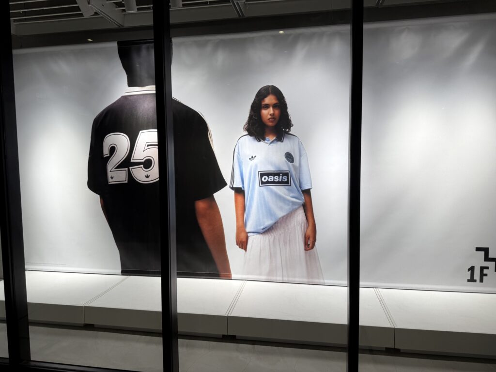 A large in-store advertisement for the Adidas and Oasis collaboration. It features a man in a black jersey with his back to the camera, and a woman facing forward wearing a light blue jersey with the Oasis logo.