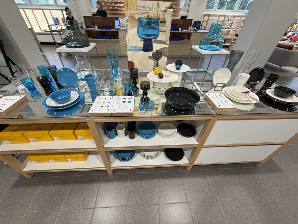 Inside a bright design center, blue, black, and white glassware and tableware are beautifully displayed on a wooden shelving unit. Yellow Iittala boxes are lined up on the bottom shelf.