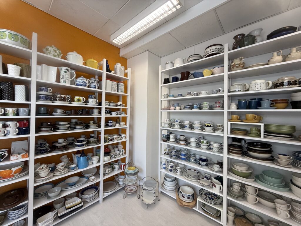 A wide variety of vintage tableware, such as cups, saucers, plates, and pots, are displayed tightly on white shelves against an orange wall.