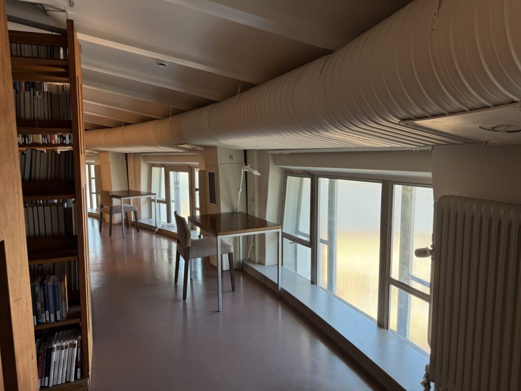 A quiet library room with study desks lined up along the windows. A large white duct runs across the ceiling, and a bookshelf is visible on the left.