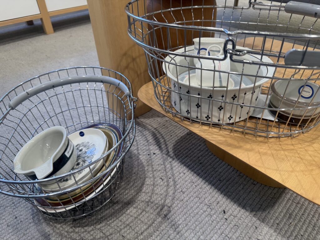 Two wire baskets, one on the floor and one on a wooden stool, filled with various vintage-style plates, bowls, and cups.