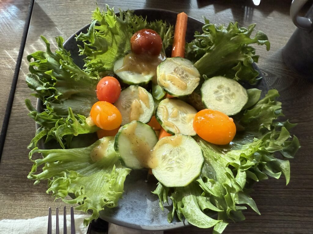 A fresh green salad with lettuce, cucumbers, cherry tomatoes, and carrots in a dark bowl on a wooden table, drizzled with dressing.