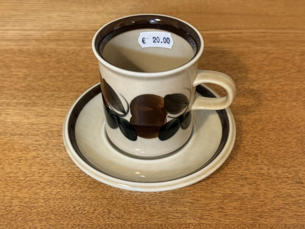 A vintage Arabia coffee cup and saucer set on a wooden table. The cup is beige with a dark brown rim and features a hand-painted style pattern of brown and black leaves. A price tag for €20.00 is visible inside the cup.
