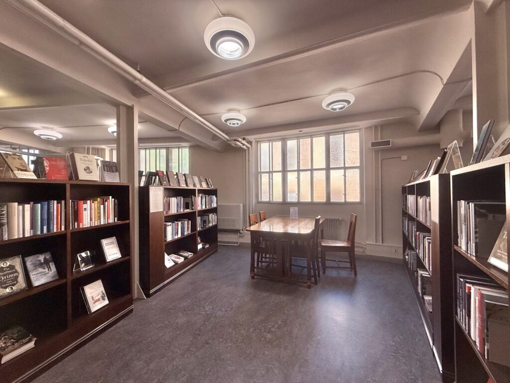 暖かい照明が灯る図書館の一室。通路の両脇に本が詰まった濃い茶色の本棚が並び、奥の窓際には複数人で使える木製のテーブルと椅子が置かれている。