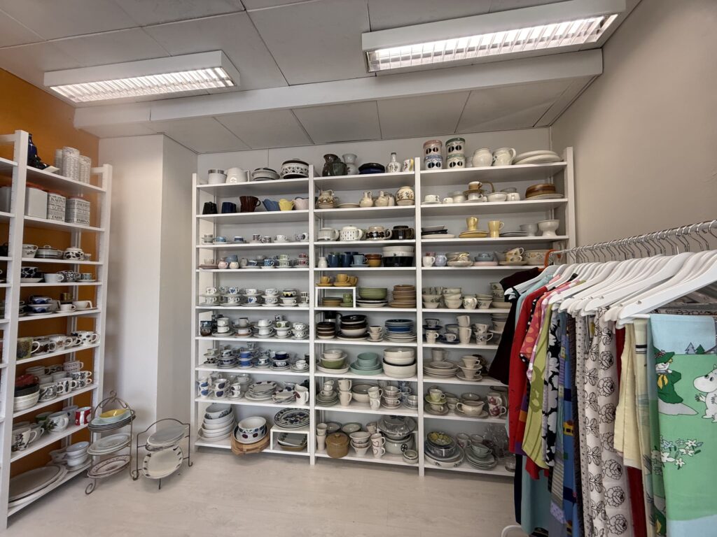 The interior of a vintage shop. White shelves are packed with Nordic design tableware such as cups, plates, and bowls, and a rack next to them holds second-hand clothes, including Moomin fabric.