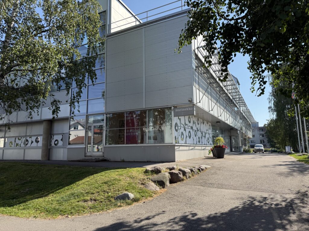 The modern exterior of the Marimekko headquarters building on a sunny day, seen past green trees. The building features large windows decorated with the iconic Unikko flower pattern.