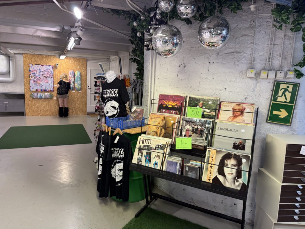 The interior of OPSHOP HELSINKI, a second-hand shop in Helsinki. In the foreground, vinyl records and graphic T-shirts are on display, and disco balls hang from the ceiling. Artworks are displayed in the background.