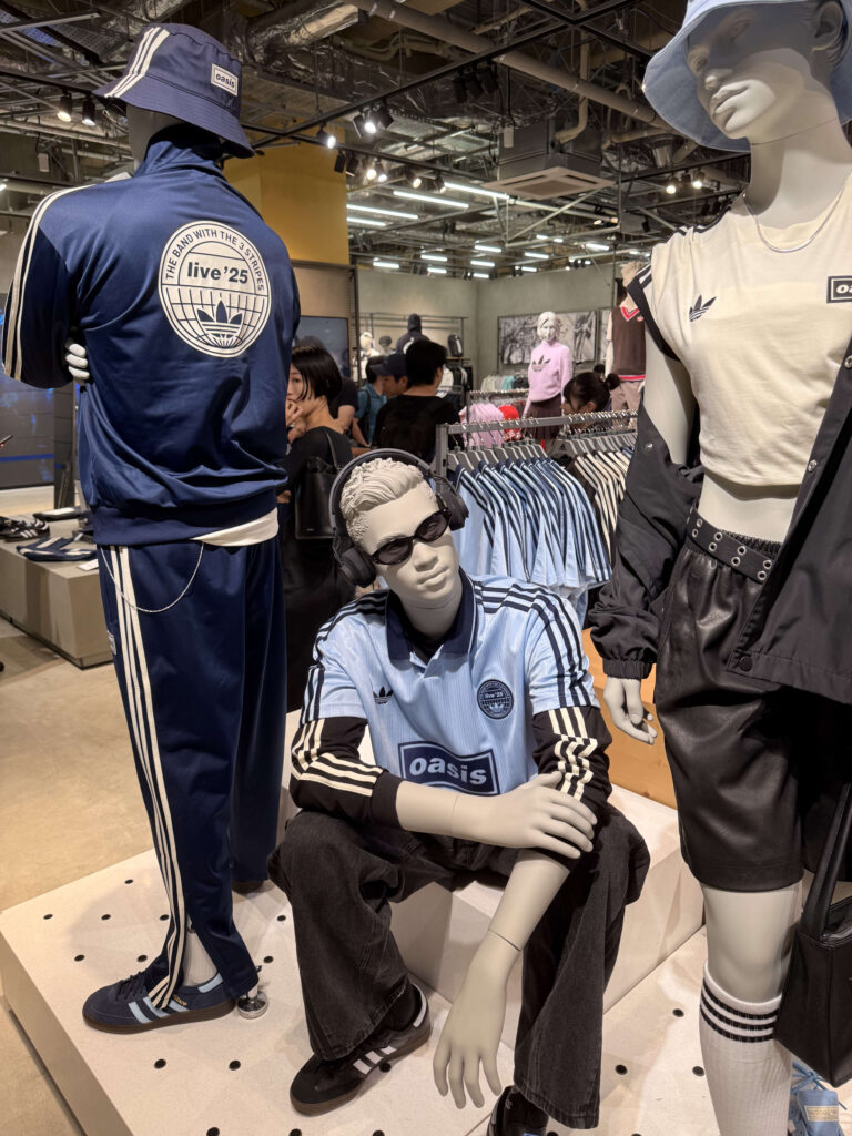 アディダスの店舗に展示された、イギリスのロックバンドOasisとのコラボレーション商品を着用した3体のマネキン。中央のマネキンは水色のサッカーシャツにサングラスとヘッドフォンを合わせ、クールに座っている。
