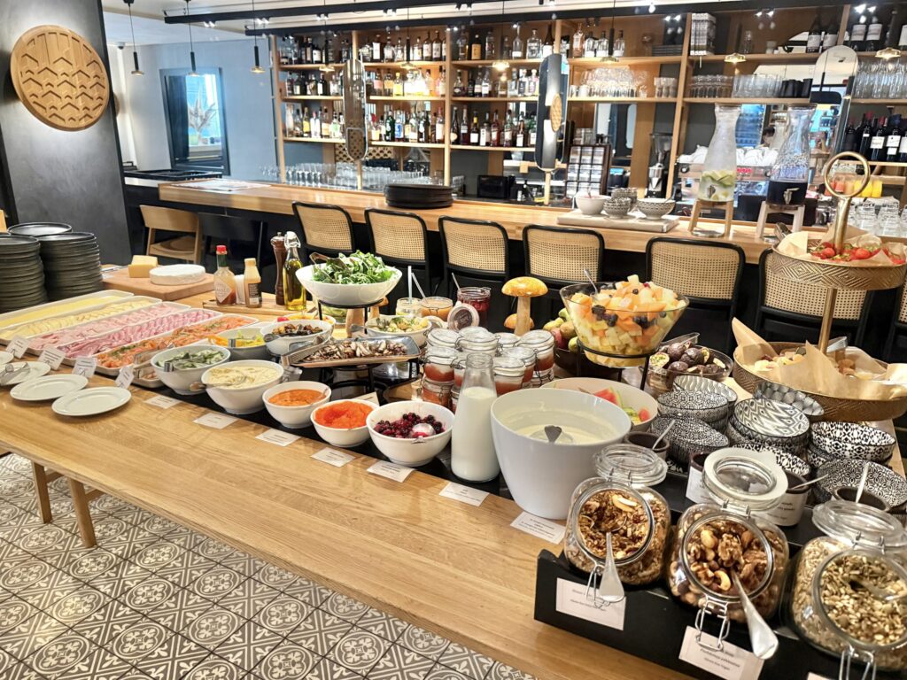 ホテルのレストランに並べられた朝食ビュッフェ。木製の長いテーブルの上に、サラダ、フルーツ、チーズ、ハムなどの色とりどりのコールドミールが並んでいる。奥にはバーカウンターが見える。