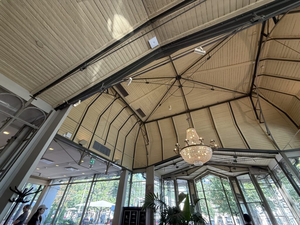 A low-angle shot looking up at the high, dome-shaped ceiling of a cafe. Light-colored wooden panels and black steel beams radiate outwards, with a large crystal chandelier hanging in the center.