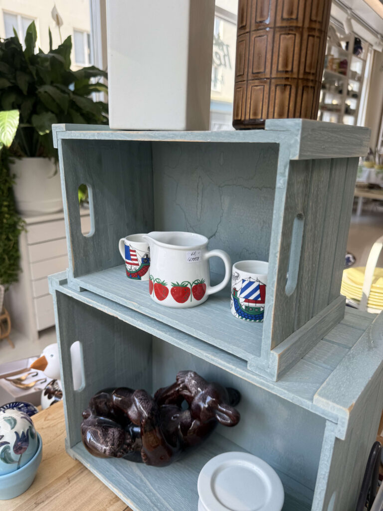 Two light-blue wooden crates are stacked, with the top one holding a pitcher with a strawberry pattern and two mugs with ship designs, and the bottom one containing a brown animal figurine.