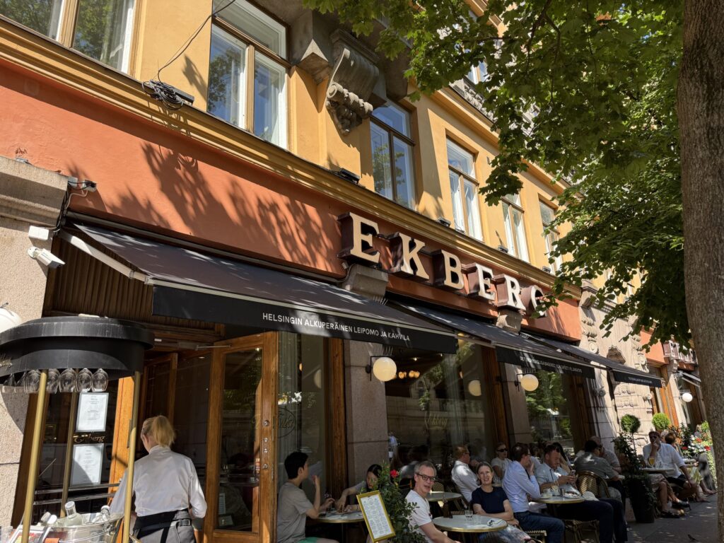 The exterior of the historic Ekberg café in Helsinki. People are relaxing at the outdoor terrace seating in front of the orange building on a sunny day.