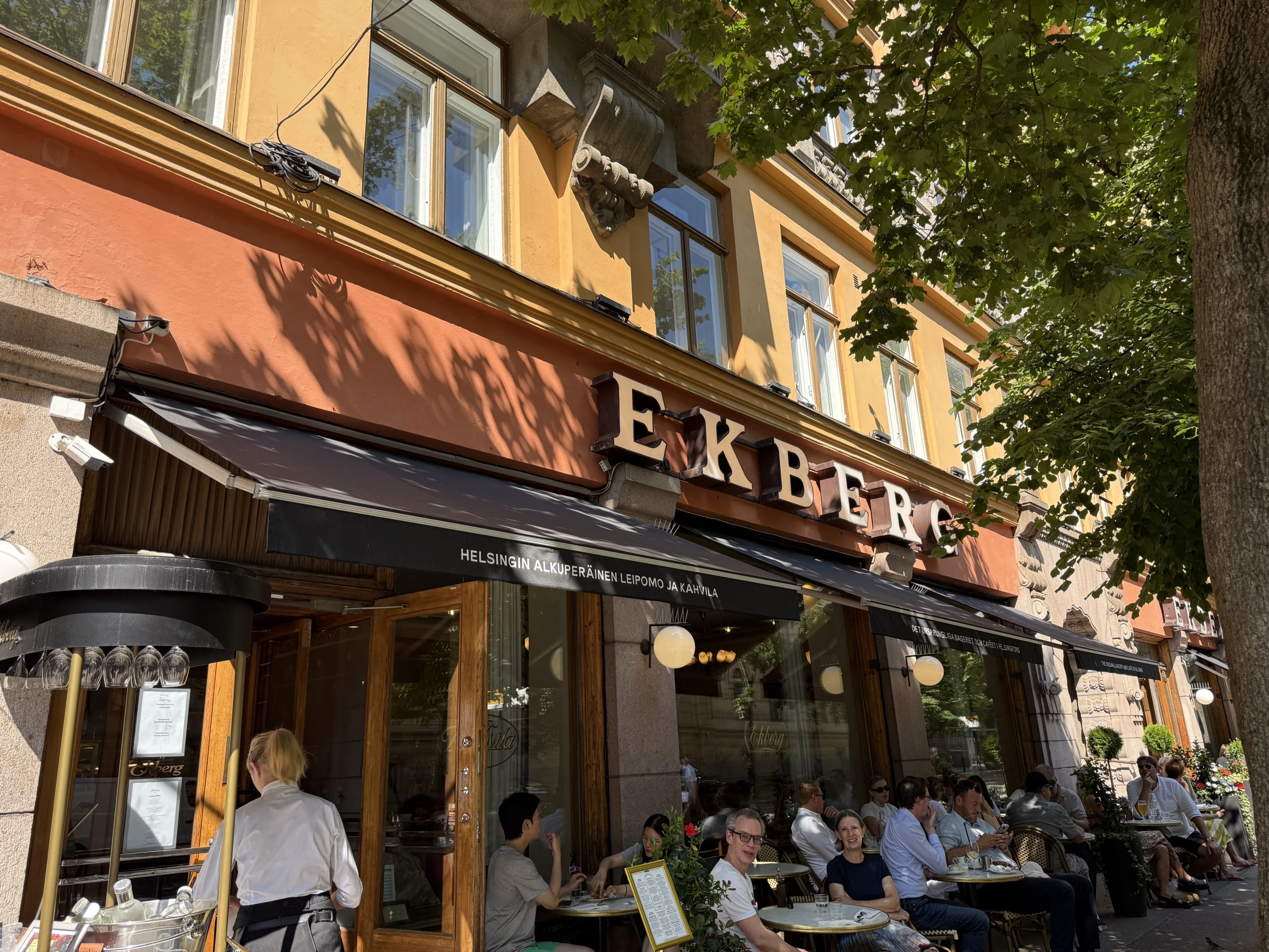The exterior of the historic Ekberg café in Helsinki. People are relaxing at the outdoor terrace seating in front of the orange building on a sunny day.