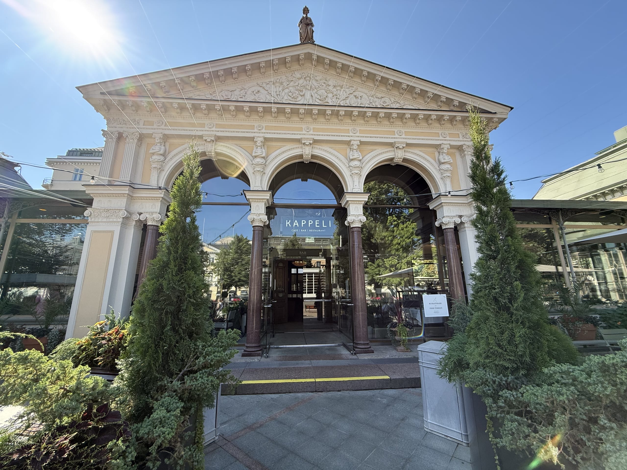 The magnificent front entrance of Restaurant Kappeli in Helsinki under a clear blue sky on a sunny day. It's a building with classical, theater-like decorations, with green trees planted on both sides of the entrance.