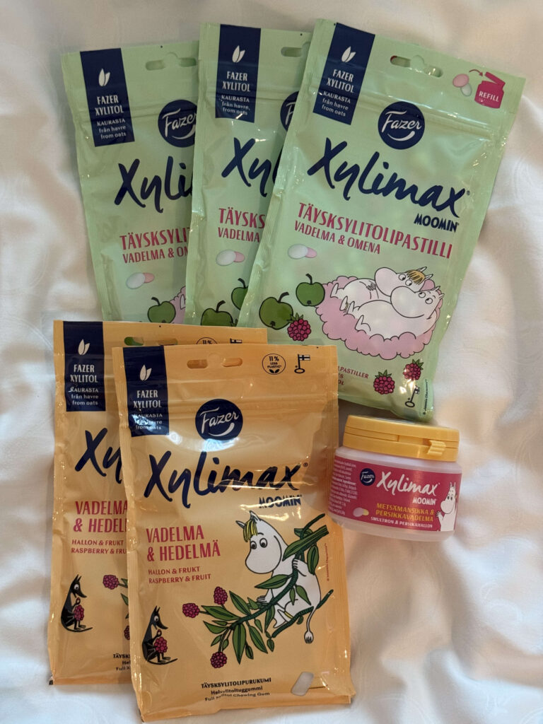 An assortment of Fazer's Moomin-themed xylitol products placed on a white cloth. There are several green and orange pouches, as well as a small plastic jar.