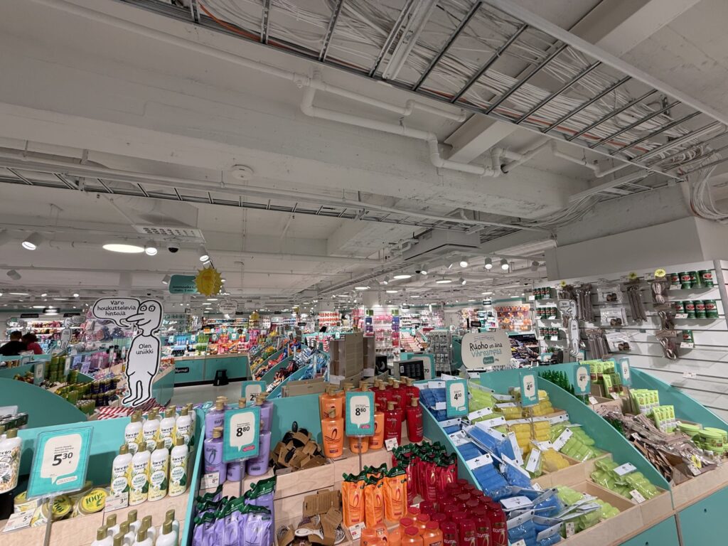 広々としたディスカウントストアの店内。ティール色の棚には、シャンプーや化粧品などのカラフルな商品がずらりと並んでいる。天井は配管がむき出しのインダストリアルなデザイン。